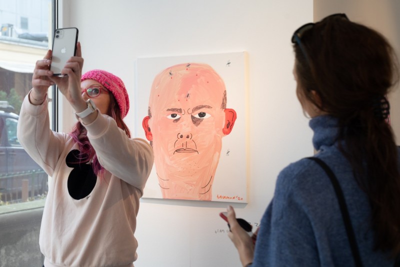 zdjęcie z przestrzeni wystawy. Na ścianie portret łysego człowieka przed obrazem dwie postacie. Jedna odwrócona w kierunku obiektywu robi autoportret telefonem, tak, żeby obraz był w kadrze. W prawej stronie zdjęcia druga osoba odwrócona do widzów plecami.