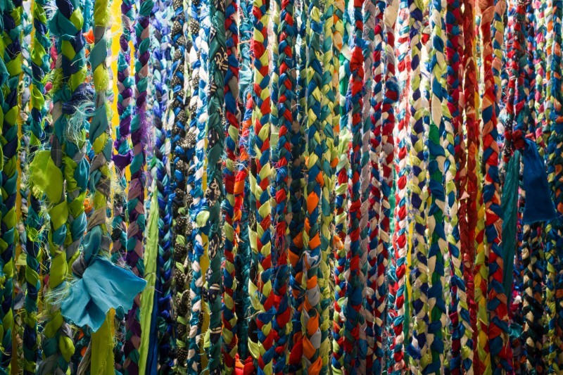 Zdjęcie przedstawiające warkocze uplecione z kolorowych ścinków materiałów. Ułożone jeden obok drugiego zapełniaja całą przestrzeń kadru